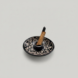 Black handmade ceramic palo santo, sage, incienso holder with white geometric patterns, containing a burning sage stick. Handame Andina World